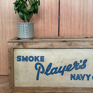 Puede incluir: Una caja de madera con un anuncio vintage de tabaco Player's Navy Cut. La caja est&aacute; pintada con las palabras "SMOKE Player's NAVY CUT" en letras azules. Una peque&ntilde;a planta verde en una maceta gris se encuentra encima de la caja.