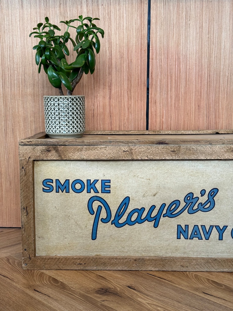 Puede incluir: Una caja de madera con un anuncio vintage de tabaco Player's Navy Cut. La caja est&aacute; pintada con las palabras "SMOKE Player's NAVY CUT" en letras azules. Una peque&ntilde;a planta verde en una maceta gris se encuentra encima de la caja.