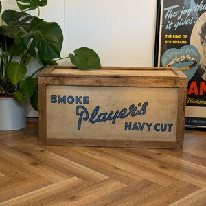 Puede incluir: Una caja de almacenamiento de madera con una etiqueta marr&oacute;n, beige y azul que dice "SMOKE Player's NAVY CUT".