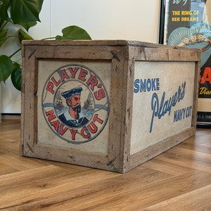 Puede incluir: Una caja de madera con un anuncio vintage de tabaco Player's Navy Cut. La caja est&aacute; pintada de marr&oacute;n y tiene una etiqueta blanca con texto azul y rojo. La etiqueta presenta a un marinero con una gorra roja y una camisa blanca. El texto de la etiqueta dice "Player's Navy Cut" y "Smoke Player's Navy Cut".