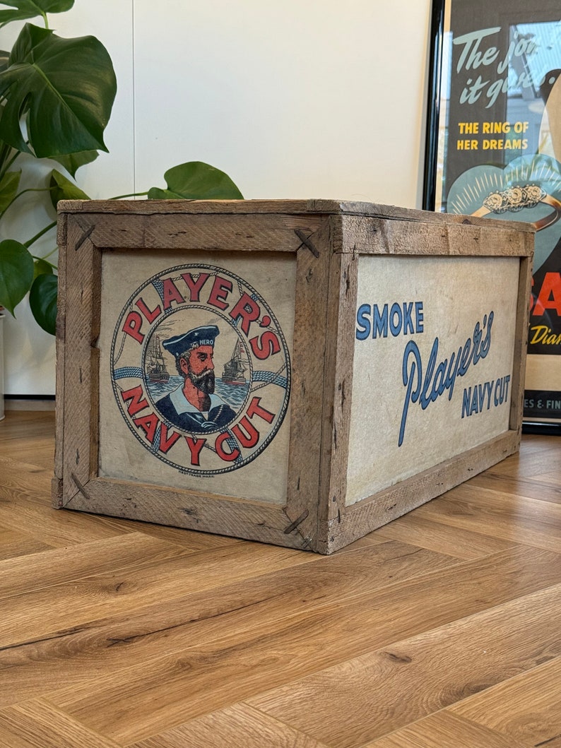 Puede incluir: Una caja de madera con un anuncio vintage de tabaco Player's Navy Cut. La caja est&aacute; pintada de marr&oacute;n y tiene una etiqueta blanca con texto azul y rojo. La etiqueta presenta a un marinero con una gorra roja y una camisa blanca. El texto de la etiqueta dice "Player's Navy Cut" y "Smoke Player's Navy Cut".