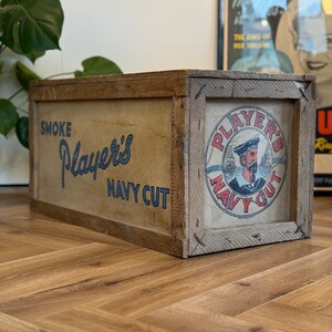 Puede incluir: Una caja de madera vintage con una etiqueta que dice "Smoke Player's Navy Cut". La etiqueta presenta a un marinero con un barco en el fondo. La caja est&aacute; sobre un suelo de madera.