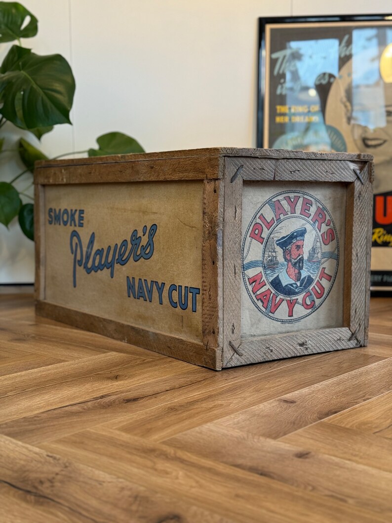 Puede incluir: Una caja de madera vintage con una etiqueta que dice "Smoke Player's Navy Cut". La etiqueta presenta a un marinero con un barco en el fondo. La caja est&aacute; sobre un suelo de madera.