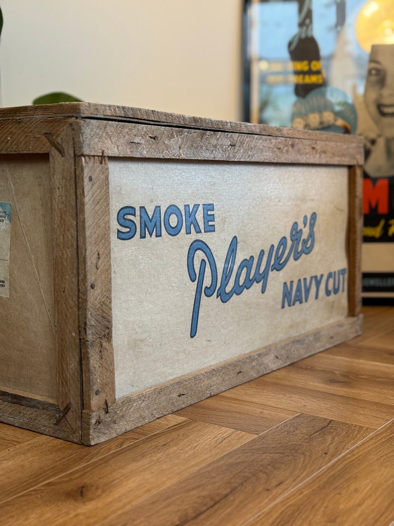 Puede incluir: Una caja de madera con una etiqueta blanca que dice "SMOKE Player's NAVY CUT" en letras azules.