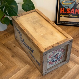 Puede incluir: Una caja de madera con una etiqueta que dice "Smoke Players Navy Cut". La caja es marr&oacute;n y tiene un aspecto desgastado. La etiqueta es azul y blanca y presenta a un marinero con un sombrero rojo.