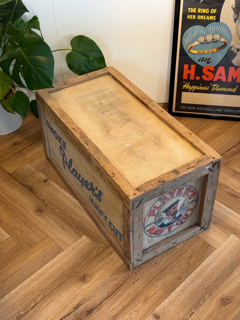 Puede incluir: Una caja de madera con una etiqueta que dice "Smoke Players Navy Cut". La caja es marr&oacute;n y tiene un aspecto desgastado. La etiqueta es azul y blanca y presenta a un marinero con un sombrero rojo.