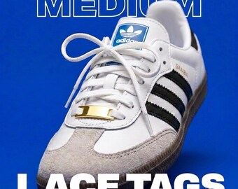May include: A white Adidas Samba trainer with black side stripes, a grey suede toe cap, and a gum rubber sole. The shoe features a gold-coloured metal lace tag on the white laces. The text MEDIUM LACE TAGS is displayed in white over a blue background.