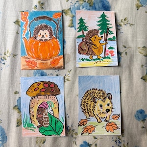 Può includere: Quattro stampe artistiche rettangolari con illustrazioni a matita colorata di ricci. Una mostra un riccio in una teiera a forma di zucca, un'altra un riccio escursionista con zaino, una terza sotto un fungo e l'ultima un riccio con foglie autunnali.