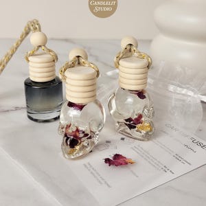 May include: Clear skull-shaped car air fresheners filled with dried rose petals and gold flakes. Each bottle has a wooden cap and a rope for hanging. The image also shows a small bottle of black liquid and a card with instructions.