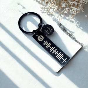 May include: Black metal keychain with a Spotify code and the initials "J+A" engraved on a round charm. The keychain includes a key ring and a small chain. The background is white with a hint of floral elements.