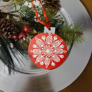 May include: Red wooden Christmas ornament with a white mandala design. The ornament is hanging from a red ribbon.