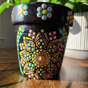 May include: A black terracotta flower pot with a hand-painted mandala design in shades of pink, green, yellow, and blue. The pot has a black rim with painted flowers in pink, green, and yellow.