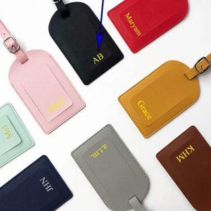 Könnte beinhalten: Satz mit neun Leder-Gepäckanhängern in verschiedenen Farben, jeder mit einem personalisierten Namen. Die Anhänger haben eine Schlaufe zum Befestigen am Gepäck.