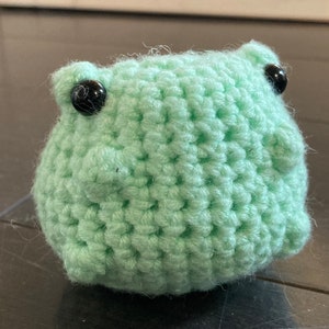 Può includere: Un peluche di rana verde all'uncinetto con occhi a bottone neri.