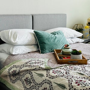 May include: A white bed with a gray headboard and a green velvet pillow. The bed is made with a white and green patterned quilt and has a wooden tray with a small plant and a decorative jar on top.
