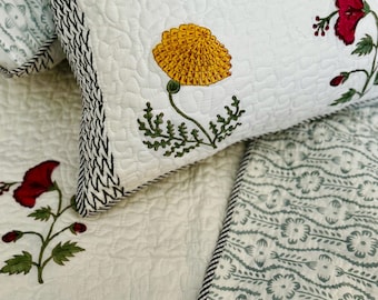 May include: White quilted pillow and bedding set with floral designs. The pillow features a yellow flower, red flower, and blue leaf pattern. The edges of the pillow have a black and white zigzag pattern. The bedding has a floral pattern with a green and white color scheme.