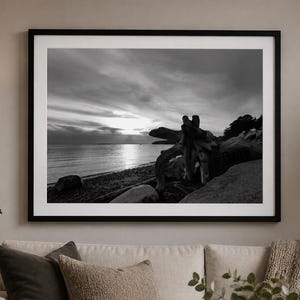 May include: A black and white landscape photograph in a thin black frame featuring a beach at sunset with a large piece of driftwood in the foreground, calm water, and a cloudy sky, displayed on a wall above a beige sofa with decorative pillows.
