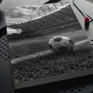 Puede incluir: Un cuaderno rectangular que presenta una fotografía en blanco y negro de un balón de fútbol descansando sobre el césped cerca de una línea lateral blanca, con un gran estadio y focos brillantes visibles al fondo bajo un cielo nublado.