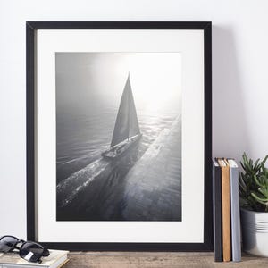 May include: Black and white framed print of a sailboat on the water. The image is in a black frame and is displayed on a wooden surface. Sunglasses and a book are in front of the frame.