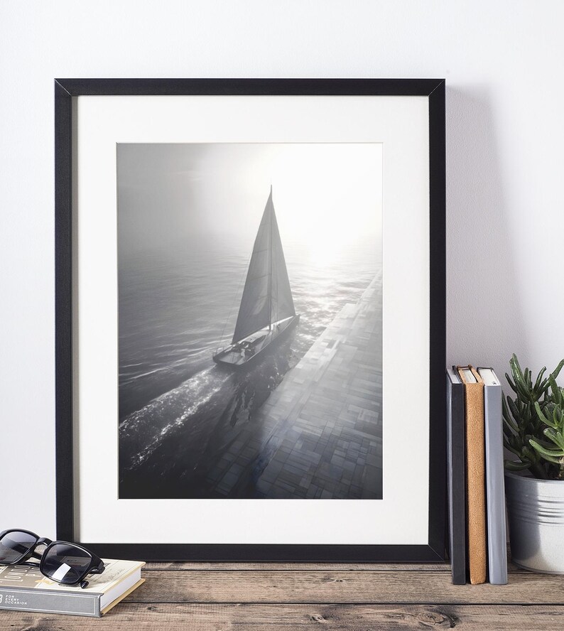 May include: Black and white framed print of a sailboat on the water. The image is in a black frame and is displayed on a wooden surface. Sunglasses and a book are in front of the frame.