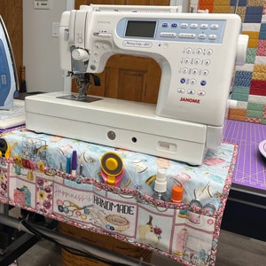 Puede incluir: Una máquina de coser blanca con una pantalla digital y una variedad de botones y diales. La máquina está sentada en una mesa con un organizador de tela que dice "Happiness is Handmade".