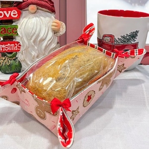 Puede incluir: Una cesta de pan de tela rosa y roja con un diseño de hombre de jengibre. La cesta está llena de una hogaza de pan y tiene una ventana de plástico transparente. La cesta está sentada sobre un mantel blanco.