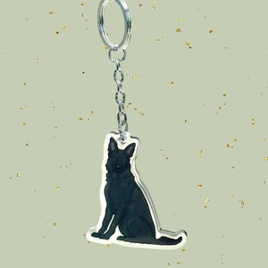 May include: A black and white illustration of a dog on a silver keychain. The dog is sitting with its head tilted to the side.