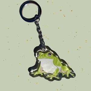 May include: A green frog-shaped keychain with a silver chain and keyring. The frog has a white belly, black eyes, and is sitting on a dark base. The background is a light green with gold flecks.