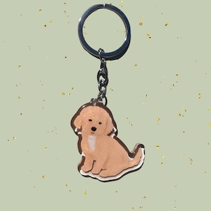May include: A keychain featuring a cartoon illustration of a golden-colored puppy. The puppy is sitting with its tail curled. The keychain has a silver-colored ring and chain. The background is a light green color with gold flecks.