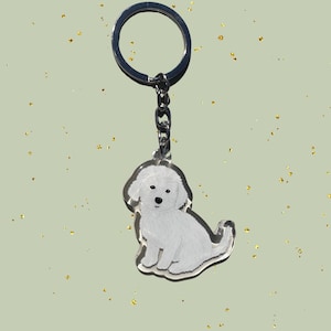 May include: A clear acrylic keychain featuring a white dog illustration. The dog is sitting with its tail curled. The keychain has a silver metal ring and chain. The background is a light green color with gold glitter.
