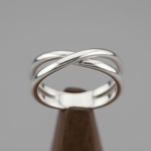 May include: A silver ring featuring a crossover design. The ring has a polished, reflective surface and a smooth, rounded form. The ring is displayed on a brown stand against a grey background. The ring appears to be made of metal.