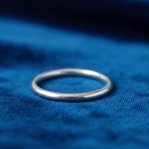 Puede incluir: Un anillo de plata con una banda lisa y redondeada. El anillo se presenta sobre un fondo azul oscuro, creando un contraste que resalta su brillo metálico. Su diseño sencillo lo hace adecuado para el uso diario.