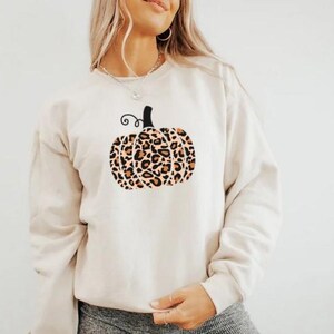 Könnte beinhalten: Ein weißer Pullover mit einem Leopardenmuster-Kürbisdesign. Der Kürbis hat einen schwarzen Stiel und einen braunen Wirbel.