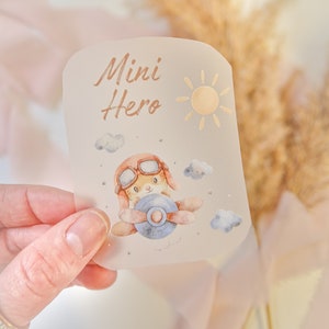Könnte beinhalten: Ein transparenter Aufkleber mit einer Aquarell-Illustration eines braunen Kaninchens, das eine Fliegerbrille trägt und ein blaues Flugzeug fliegt. Der Aufkleber trägt die Aufschrift "Mini Hero" in goldenen Buchstaben und enthält eine goldene Sonnenillustration.