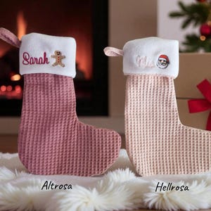 Könnte beinhalten: Zwei Weihnachtsstrümpfe, einer in Staubrosa und der andere in Hellrosa, mit weißen Bündchen. Der staubrosafarbene Strumpf hat den Namen "Sarah" und einen Lebkuchenmann. Der hellrosa Strumpf hat den Namen "Miles" und ein Santa-Gesicht.