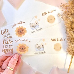 Könnte beinhalten: Drei transparente Lesezeichen mit niedlichen Illustrationen und Text. Das erste Lesezeichen zeigt eine Sonnenblume mit dem Text "Cute like my mom" und "Smile and Shine". Das zweite Lesezeichen zeigt eine Kamera mit dem Text "kleiner Entdecker" und "Little Sunshine". Das dritte Lesezeichen zeigt eine Sonne mit dem Text "Little Sunshine".