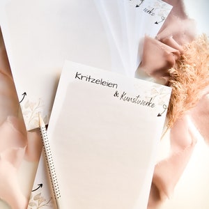 Könnte beinhalten: Ein Stapel leerer Notizblöcke mit einem floralen Design und dem Text "Kritzeleien & Kunstwerke" auf dem oberen Notizblock. Die Notizblöcke liegen auf einer weißen Oberfläche mit einem rosa und beigefarbenen Stoff.