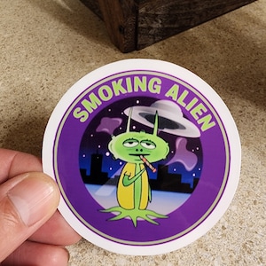 May include: A round sticker with a purple background and a white border. The sticker features a green alien smoking a cigarette in front of a city skyline. The text "SMOKING ALIEN" is written in a white circle around the alien.