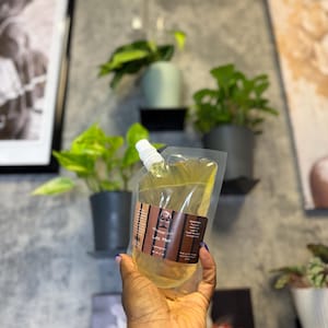 May include: A clear plastic pouch filled with a yellow liquid, labeled "Loc Mist." The pouch has a white spout and a brown label with text and a small illustration. The background includes potted plants and framed artwork.