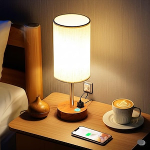 May include: A bedside table lamp with a cylindrical white lampshade and a wooden base. The lamp has a metal stem and a built-in USB charging port. A smartphone is charging on the table, next to a cup of coffee.