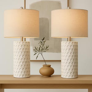 May include: Two matching cream-colored table lamps with beige fabric shades. The lamp bases are cylindrical with a repeating arched pattern. A small brown vase with a sprig of greenery sits between the lamps on a light wood surface.