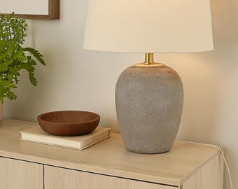 May include: A gray ceramic table lamp with a cream-colored lampshade. The lamp has a brass-colored neck and finial. A wooden bowl sits on a book next to the lamp. The lamp is on a light wood cabinet.
