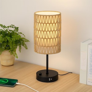 May include: A cylindrical table lamp with a woven beige shade and a black base. The shade has a geometric pattern, and the base includes a power button and USB ports. The lamp is plugged in and sits on a wooden surface.