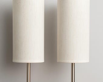May include: Two matching table lamps with cylindrical off-white fabric shades. The lamps have a brushed metal stem and sit on a round, light-colored wooden base with a black power outlet. The lamps are on a wooden surface.