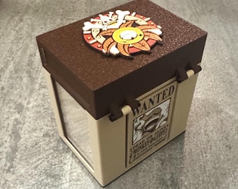 May include: A rectangular storage box with a dark brown textured lid and beige sides. The lid features a circular emblem with a skull and crossbones. One side displays a rectangular poster with the text WANTED DEAD OR ALIVE MONKEY D LUFFY.