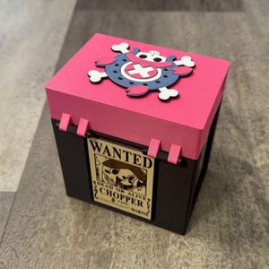 May include: A black and pink box with a pink lid featuring a blue and white skull and crossbones design. The box has a "Wanted" poster with the text "Chopper" on the front.