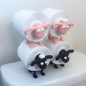 May include: Two white toilet paper rolls are held by two pink sheep figurines, and two more white toilet paper rolls are held by two black sheep figurines. The sheep figurines are holding the toilet paper rolls in their mouths.