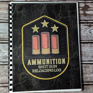 May include: A spiral-bound notebook with a black leather-like cover. The cover features a gold and black emblem with three stars and three red and tan shotgun shell illustrations. The text "AMMUNITION SHOT GUN RELOADING LOG" is printed below the illustrations.