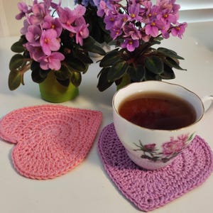 May include: A cup of tea sits on a heart-shaped, lavender crochet coaster. A matching pink crochet coaster is next to it. Two potted African violets with pink and purple flowers are in the background. The cup has a floral design.