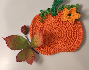 Könnte beinhalten: Ein runder, orangefarbener Häkel-Untersetzer in Kürbisform mit grünem Stiel, geschwungenen grünen Ranken und einer kleinen gelben Blüte oben. Daneben liegt eine einzelne Brombeere an einem Stiel mit drei gezackten, rot-grünen Blättern.